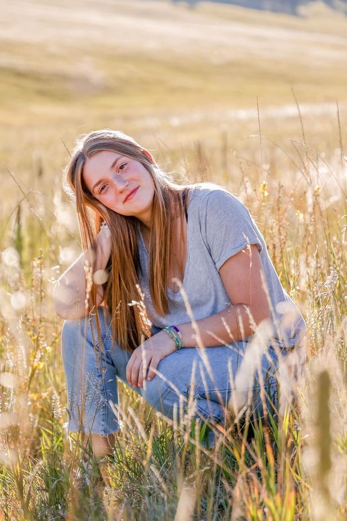Senior kneeling in the tall meadow grass, smiling with her head tilted, at Elk Meadow