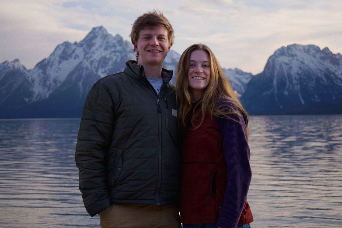 Couple standing close and smiling on the lakeshore with the snow-capped Tetons behind them