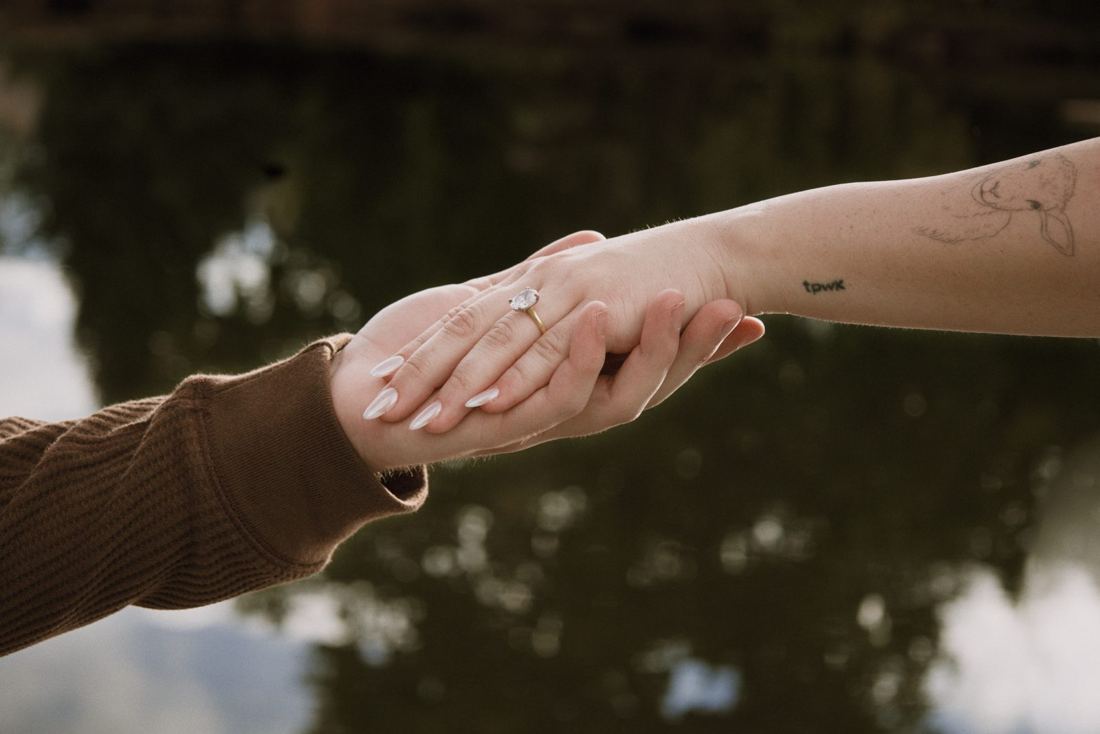 Close-up of her hand and engagement ring in his, with the pond reflecting the sky below