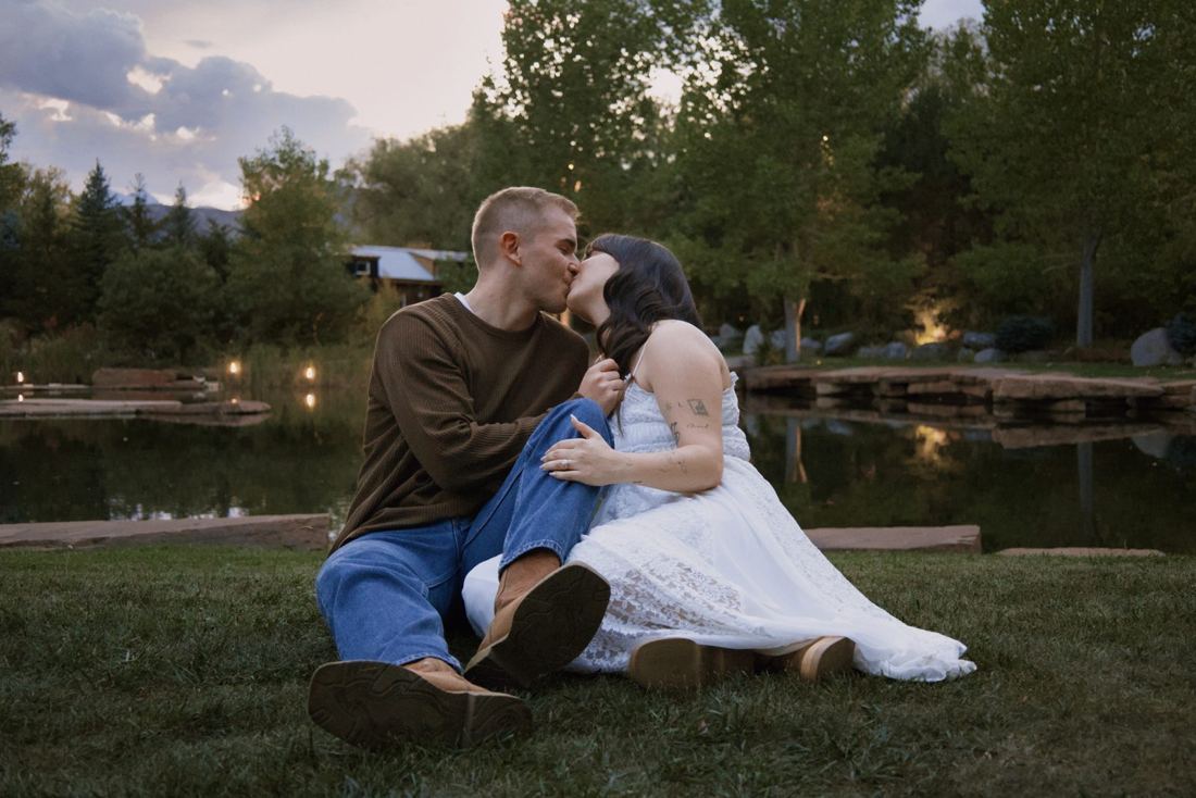 Couple sitting in the grass sharing a kiss as the string lights come on across the pond
