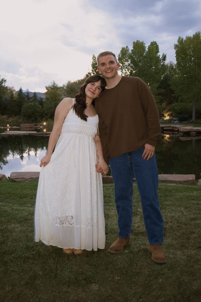 Couple standing hand in hand by the pond at dusk with string lights glowing behind them