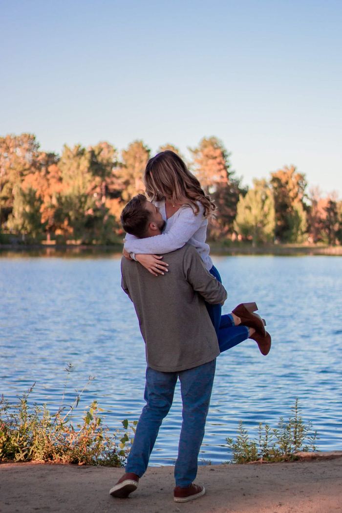 He lifts her for a kiss with the lake and fall trees behind them in Washington Park