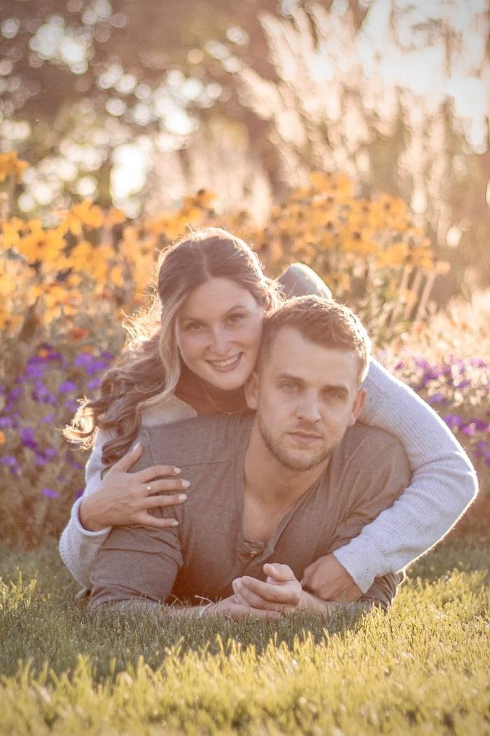 Couple lying in the grass with her arms around him in front of the flower gardens