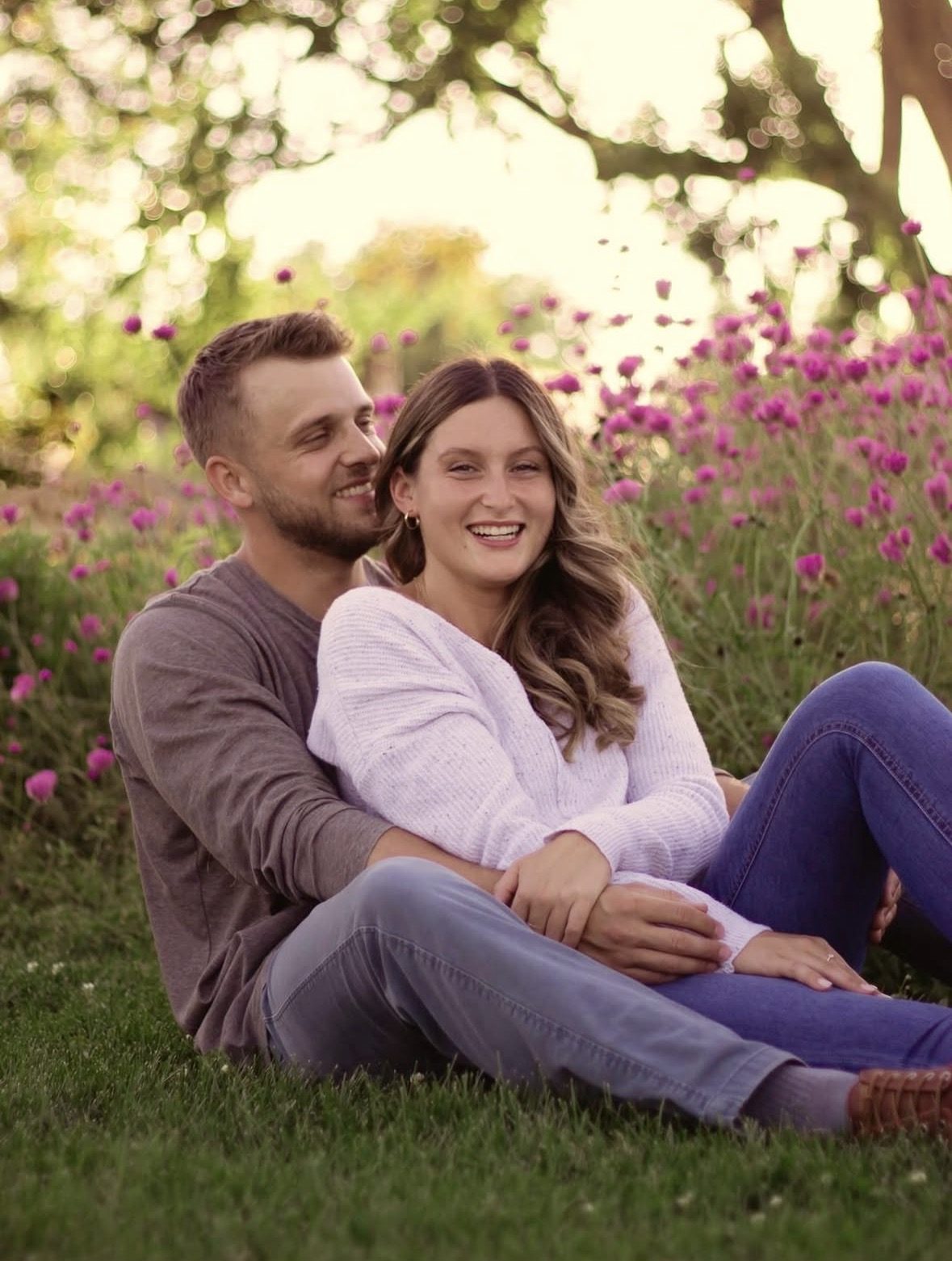 Couple sitting in the grass in front of pink flowers, both smiling at golden hour in Wash Park